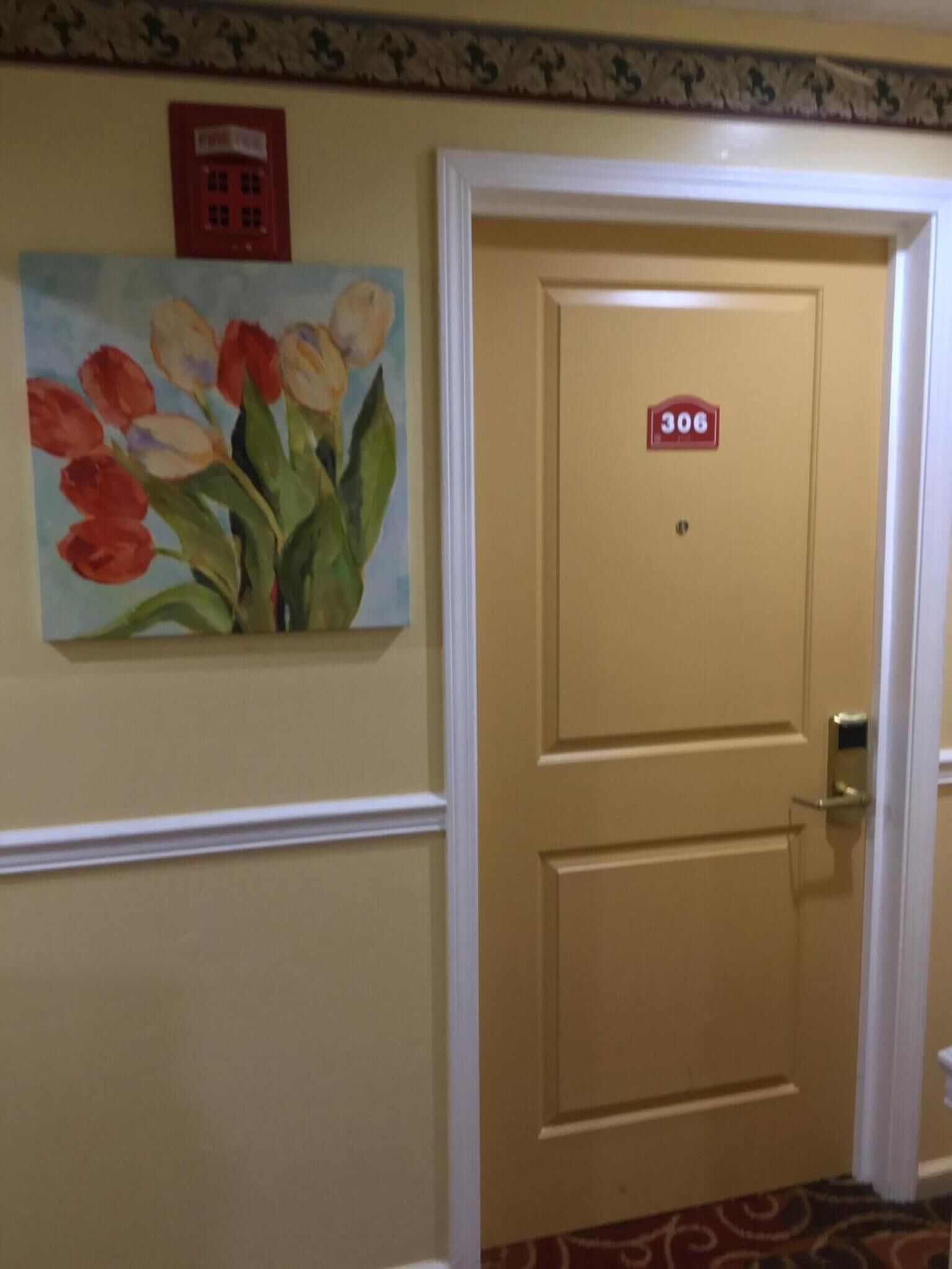 Habitación de hotel con una puerta color beige numerada 306, un teléfono rojo de emergencia y un cuadro de tulipanes en la pared.