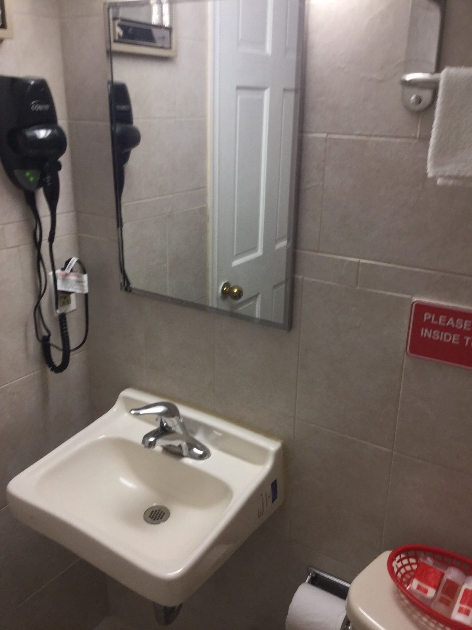 A bathroom with a sink, a mirror, a hairdryer, and a sign that says "PLEASE INSIDE TOILET".