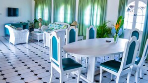 A dining area with a round table and white chairs with teal cushions, surrounded by green curtains and a television on the wall.
