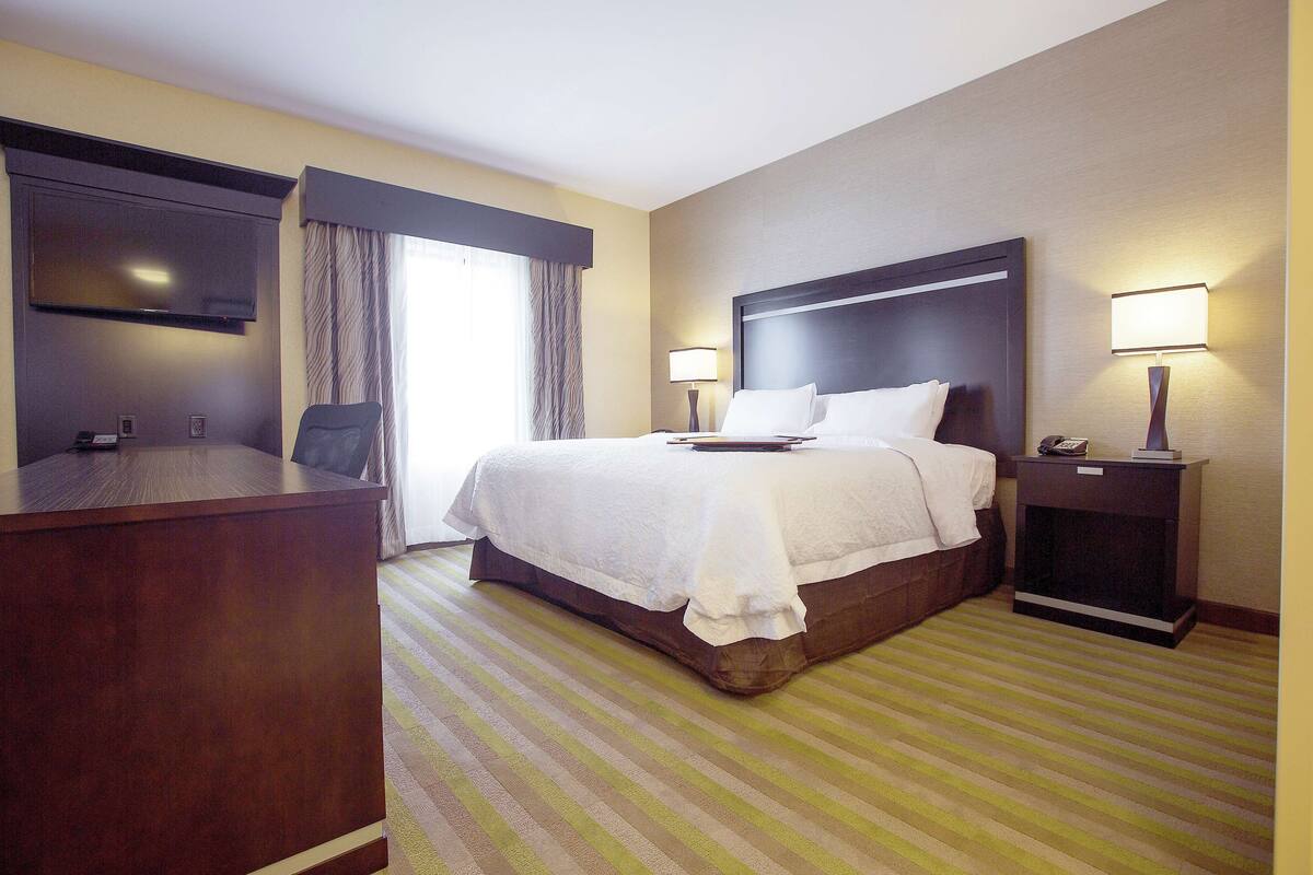 Quarto de hotel com uma cama grande, uma escrivaninha, uma televisão e uma mesa de cabeceira com abajur.