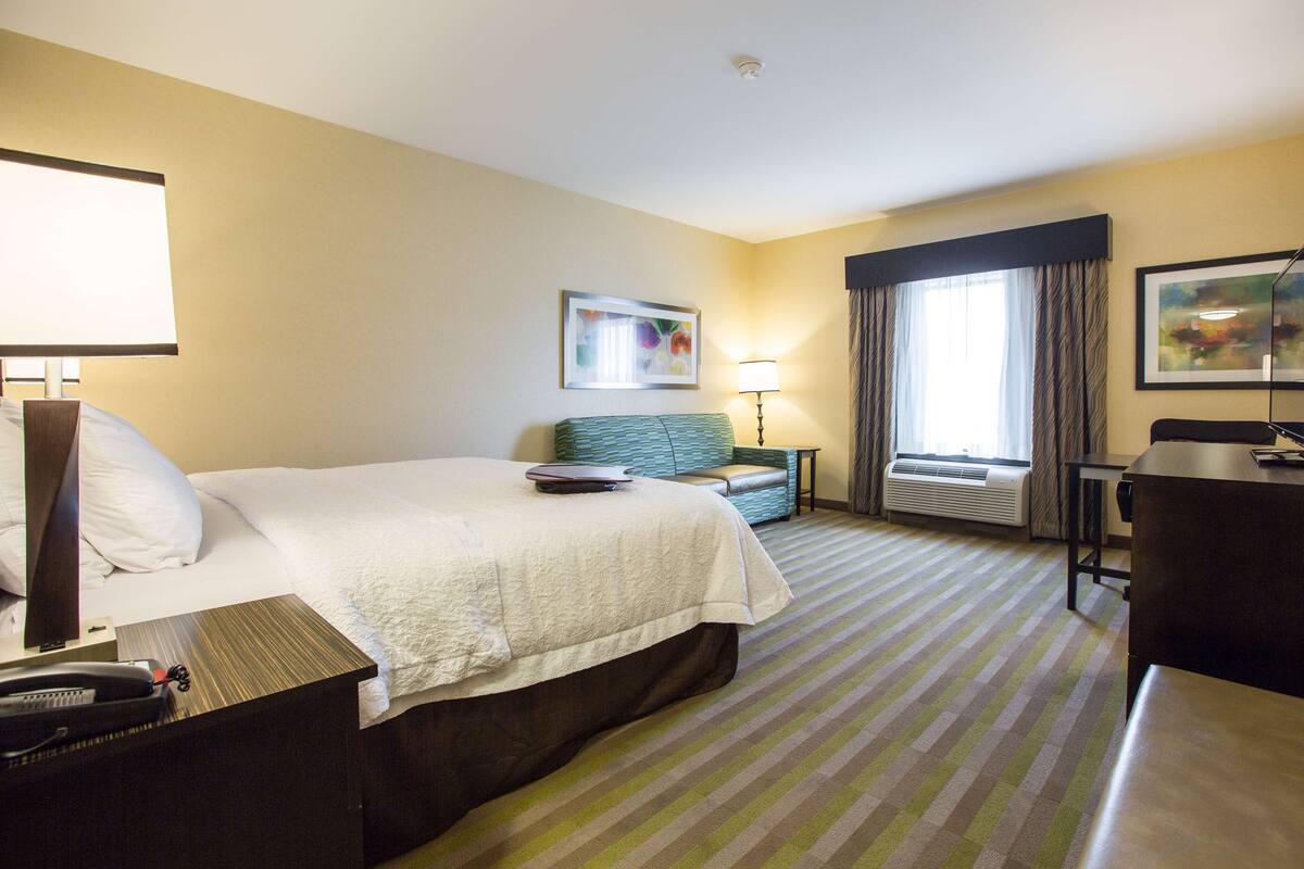 Quarto de hotel com cama, mesa de cabeceira, luminária, poltrona e escrivaninha com cadeira.