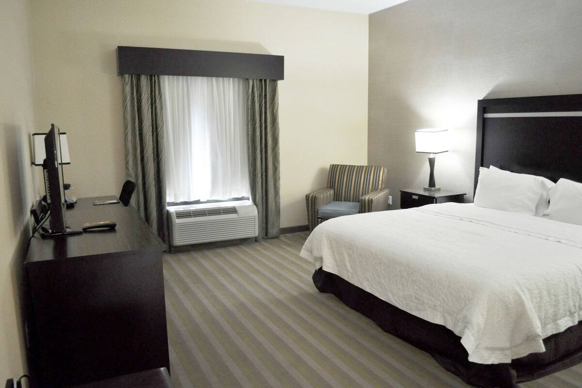 Quarto de hotel com uma cama grande, uma mesa com telefone, uma cadeira, um abajur e uma janela com cortinas.