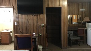 Habitación de hotel con paneles de madera, un televisor montado en la pared, un escritorio con una silla, una cama con ropa de cama a rayas y una pequeña zona de kitchenette.