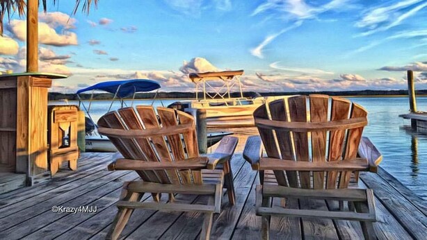Een houten steiger met twee houten stoelen en een tafel, met uitzicht op kalm water en een verre oever onder een licht bewolkte lucht.