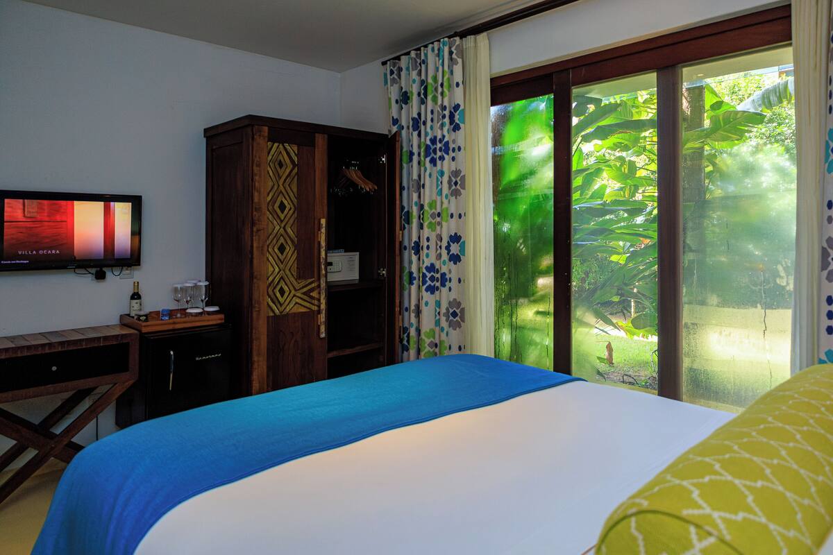 A hotel room with a bed, a desk with a TV, a minibar, and a view of greenery through the window.