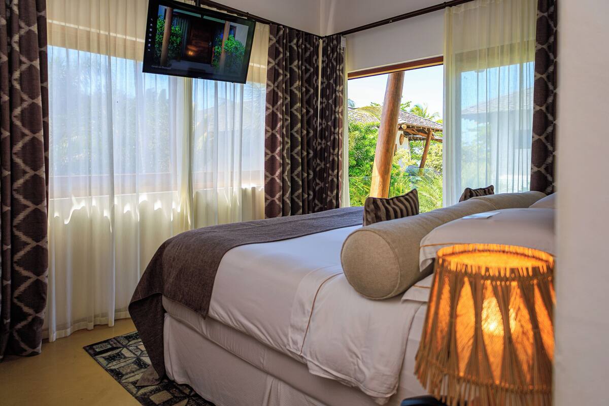 A hotel room with a bed, a TV mounted on the wall, and a view of the outdoors through large windows.
