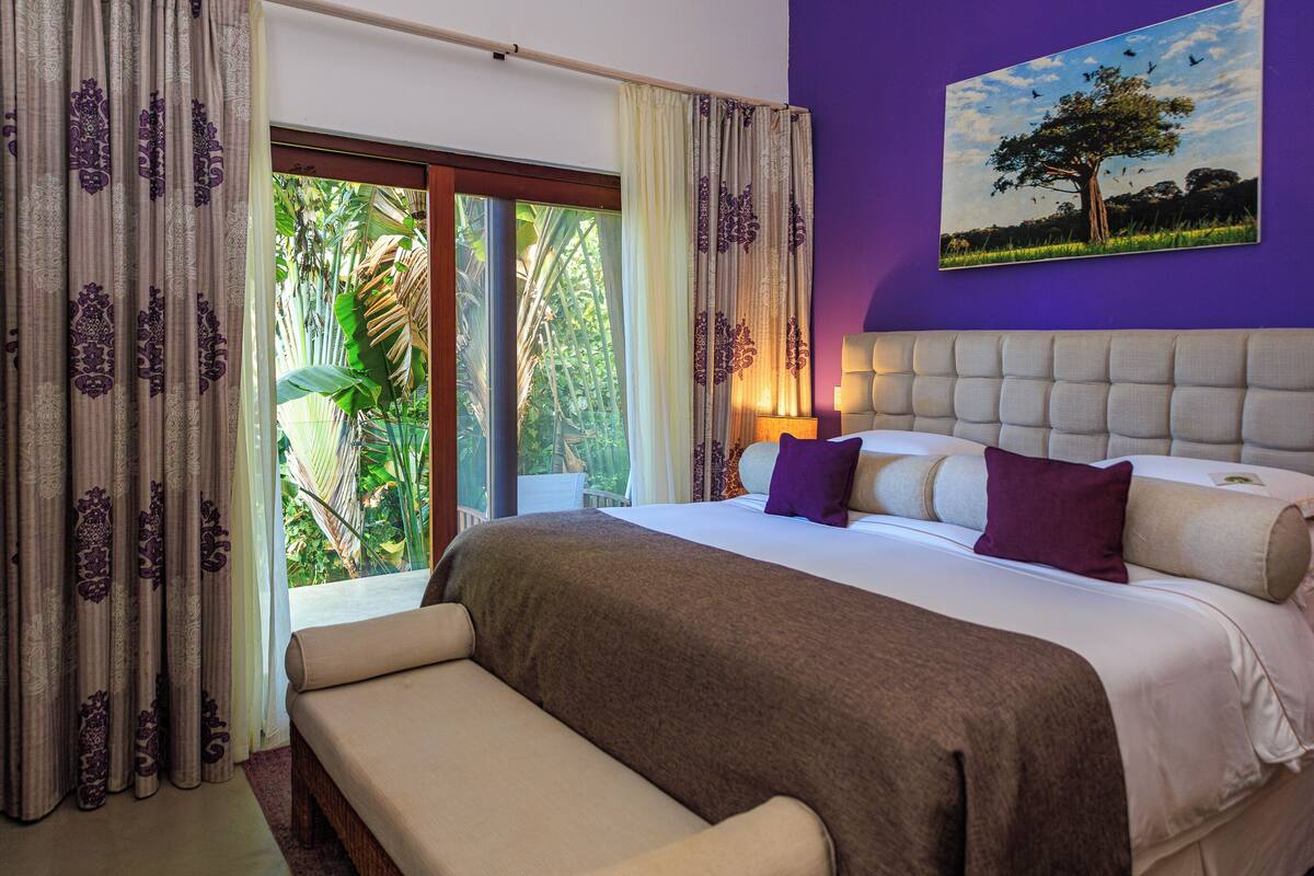A bedroom with a large bed, a sofa, and a view of greenery through the window.