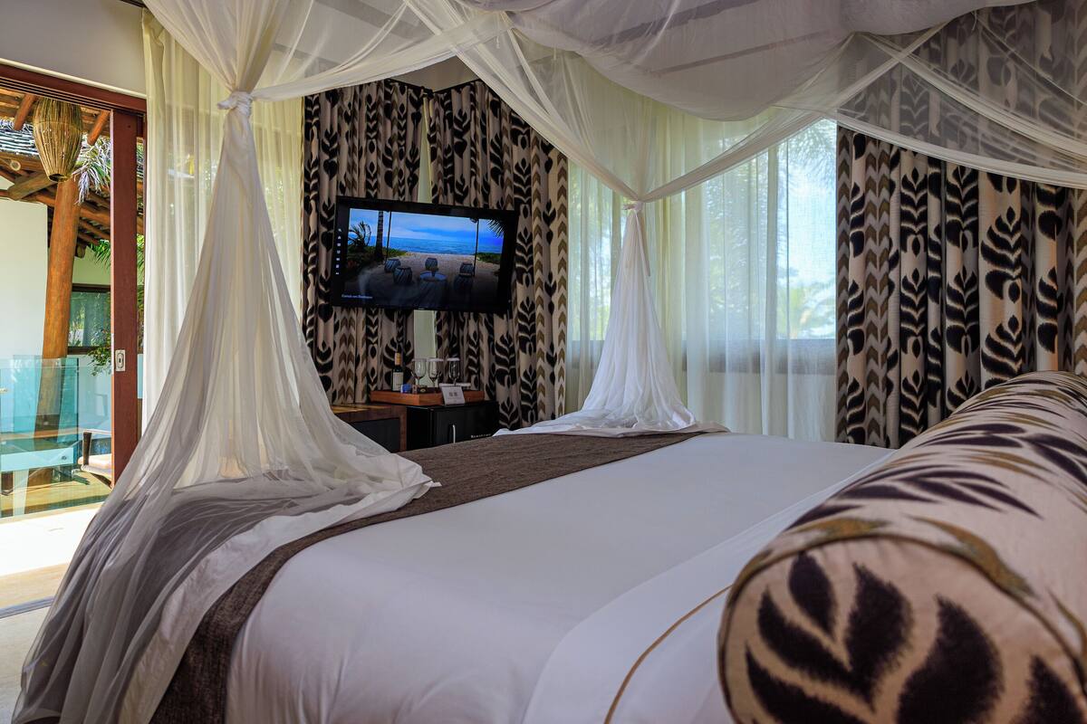 A bedroom with a large bed, a TV mounted on the wall, and sheer curtains.