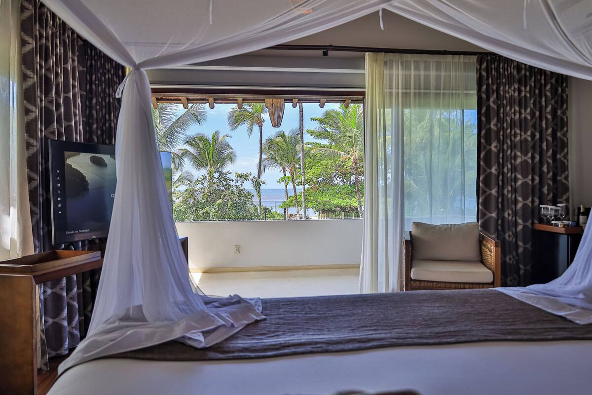 A bedroom with a bed, a television, a chair, and a view of palm trees and the ocean.