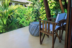 Une chaise en bois et un tabouret en osier sur une terrasse en béton, le tout entouré d’une végétation luxuriante.