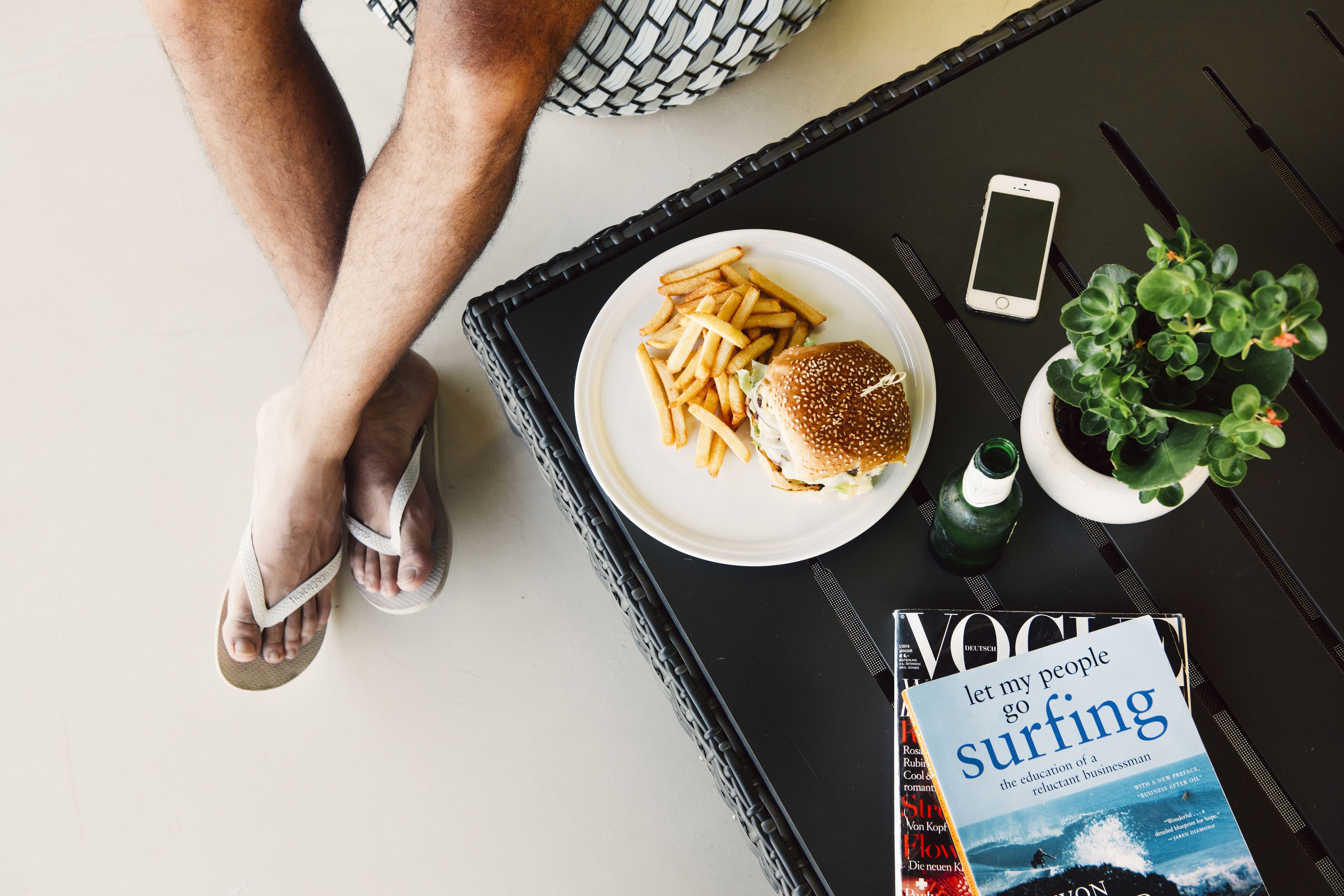 Uma bandeja com um hambúrguer e batatas fritas, um smartphone, uma revista e uma pequena planta sobre uma mesa.