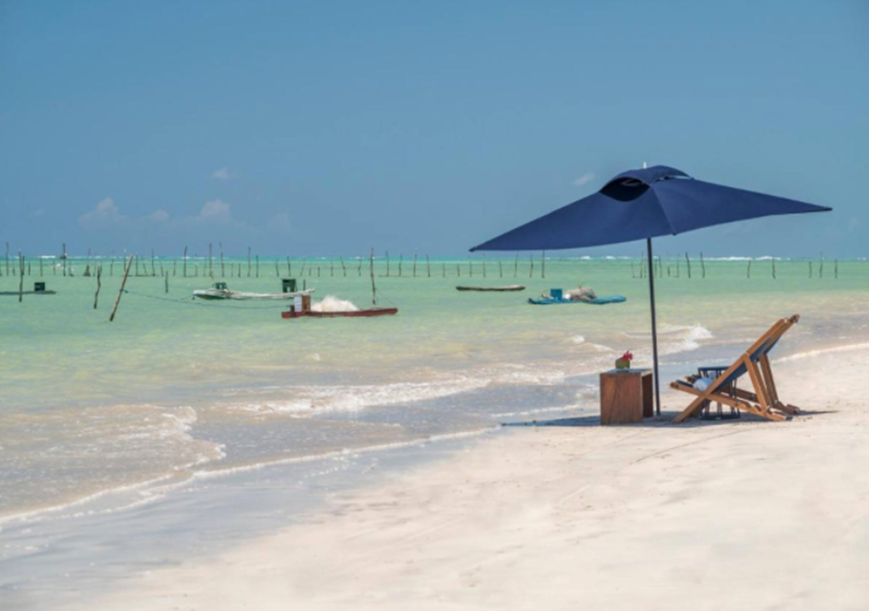 A beach with a blue umbrella, a wooden chair, and a small wooden table.