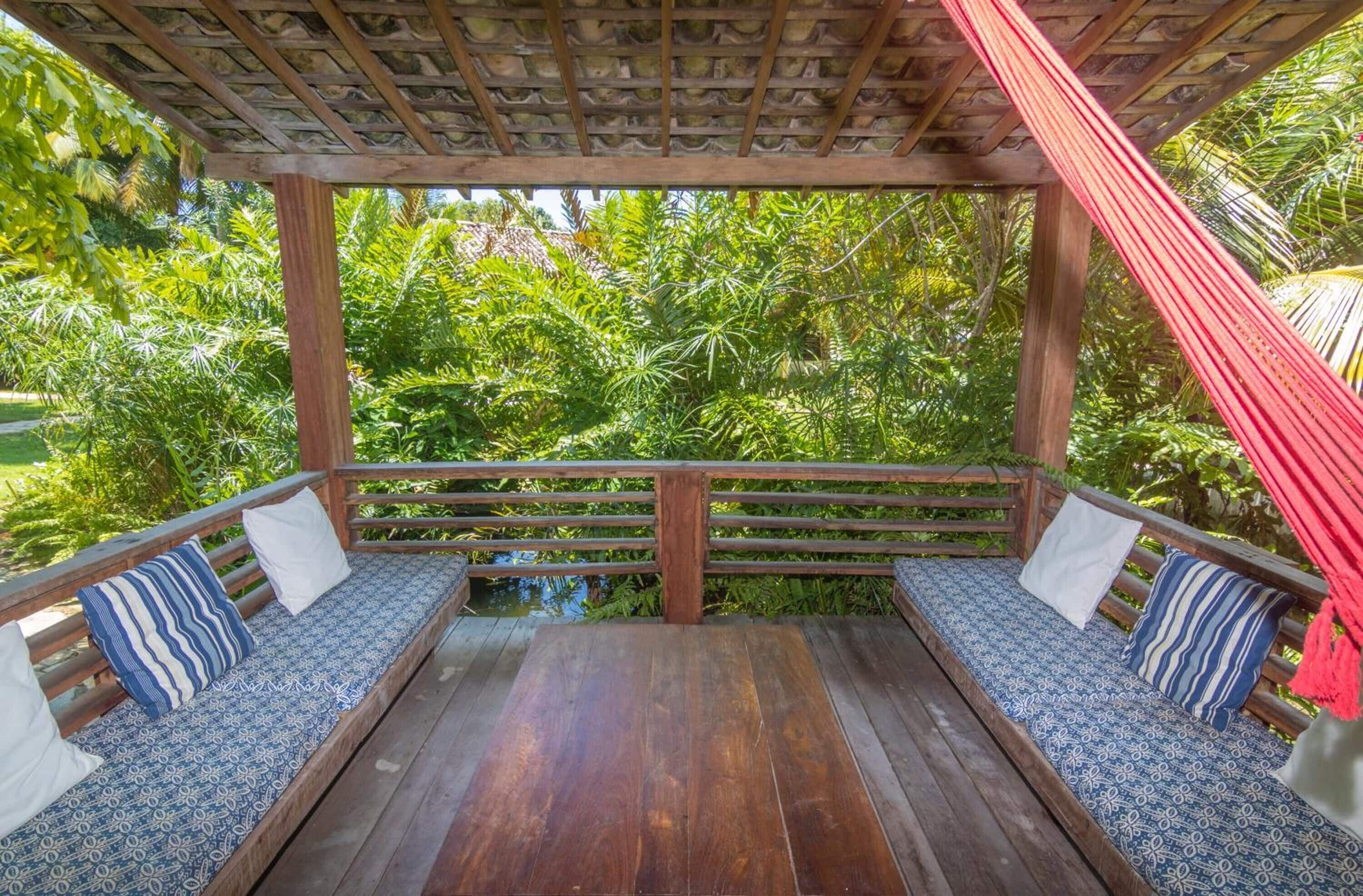 A wooden deck with a hammock, two benches with patterned cushions, and a view of lush greenery.