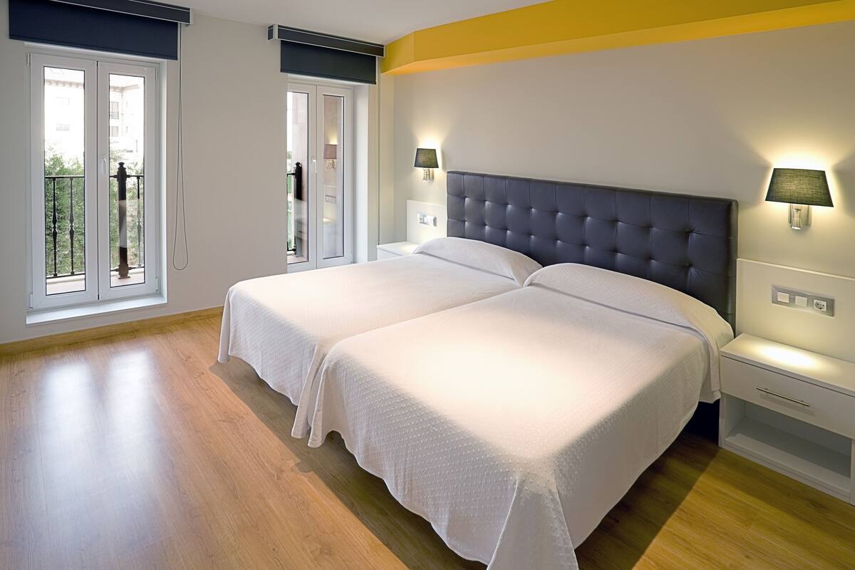 A double bed with white linens, two bedside tables with lamps, and a view of a balcony through a glass door.