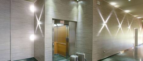 A modern hotel lobby with a glass elevator, a reception desk, and two suitcases on the floor.