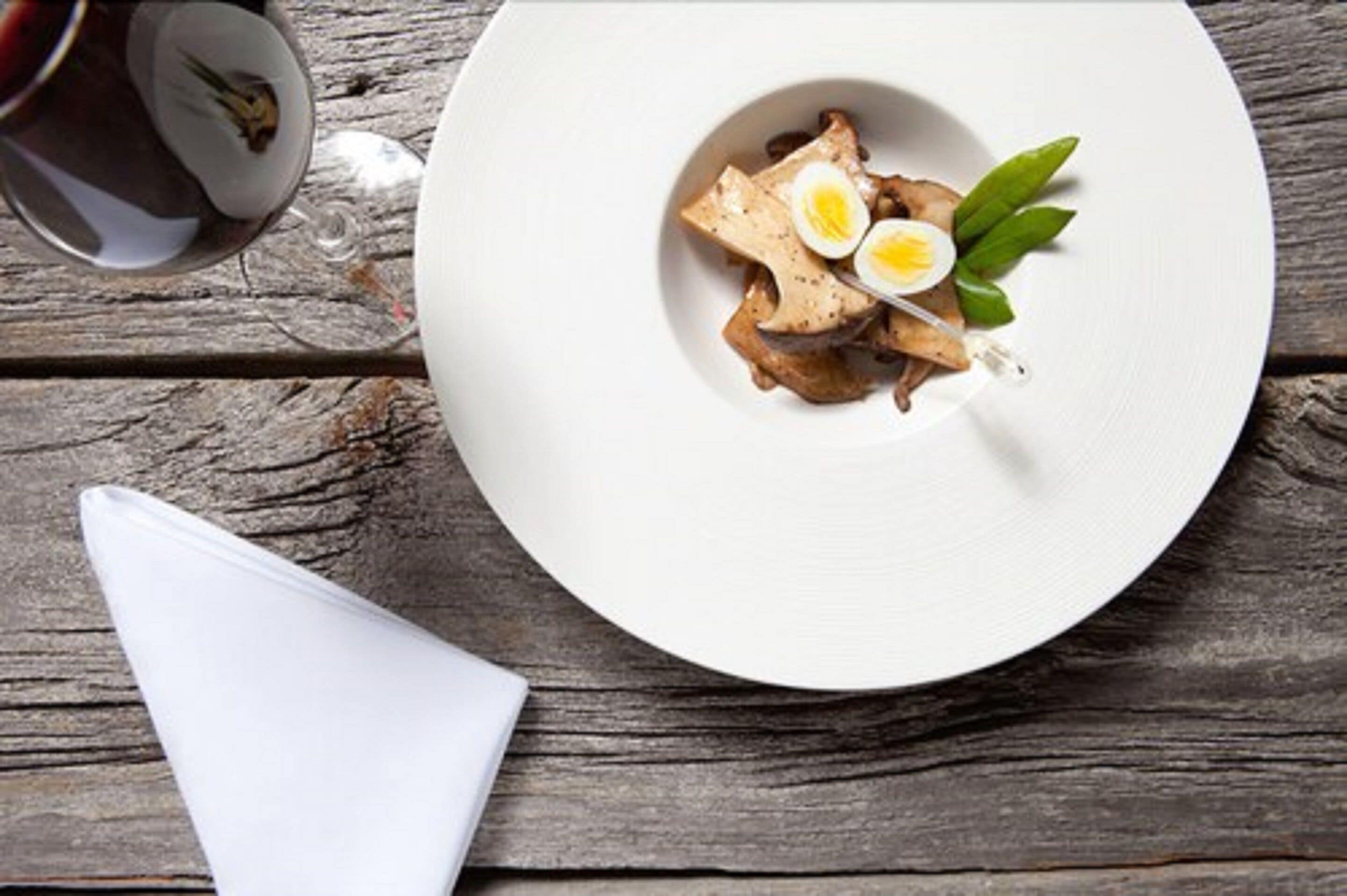 A white plate with a dish of sliced meat, a boiled egg, and green leaves on a wooden table.