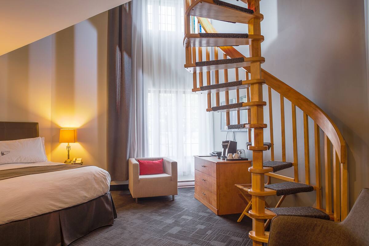 Ein Hotelzimmer mit Bett, Schreibtisch, Stuhl, einer Holztreppe und einem fenster mit Vorhängen.