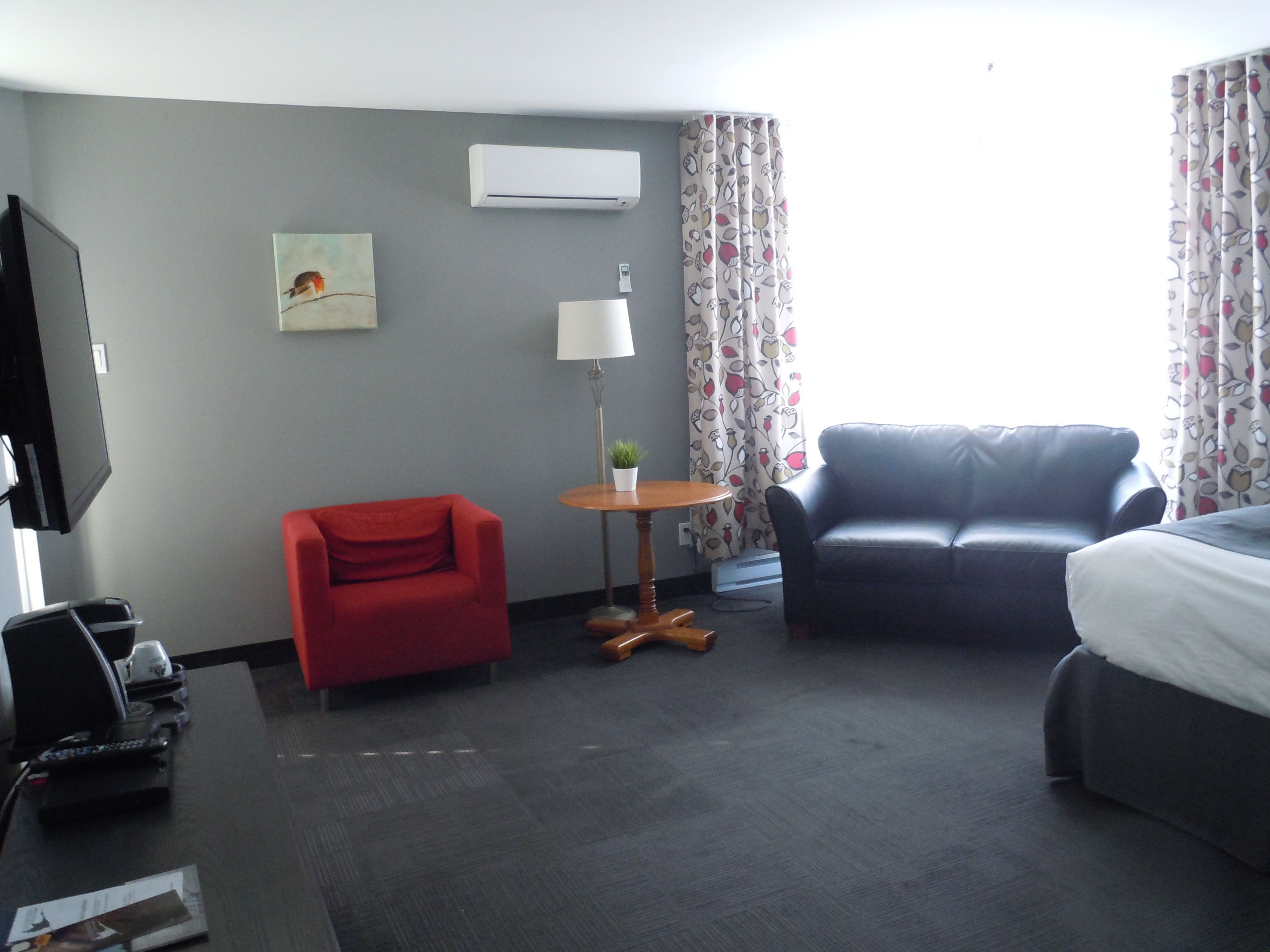 A hotel room with a red armchair, a black sofa, a small round table with a lamp, and a bed.