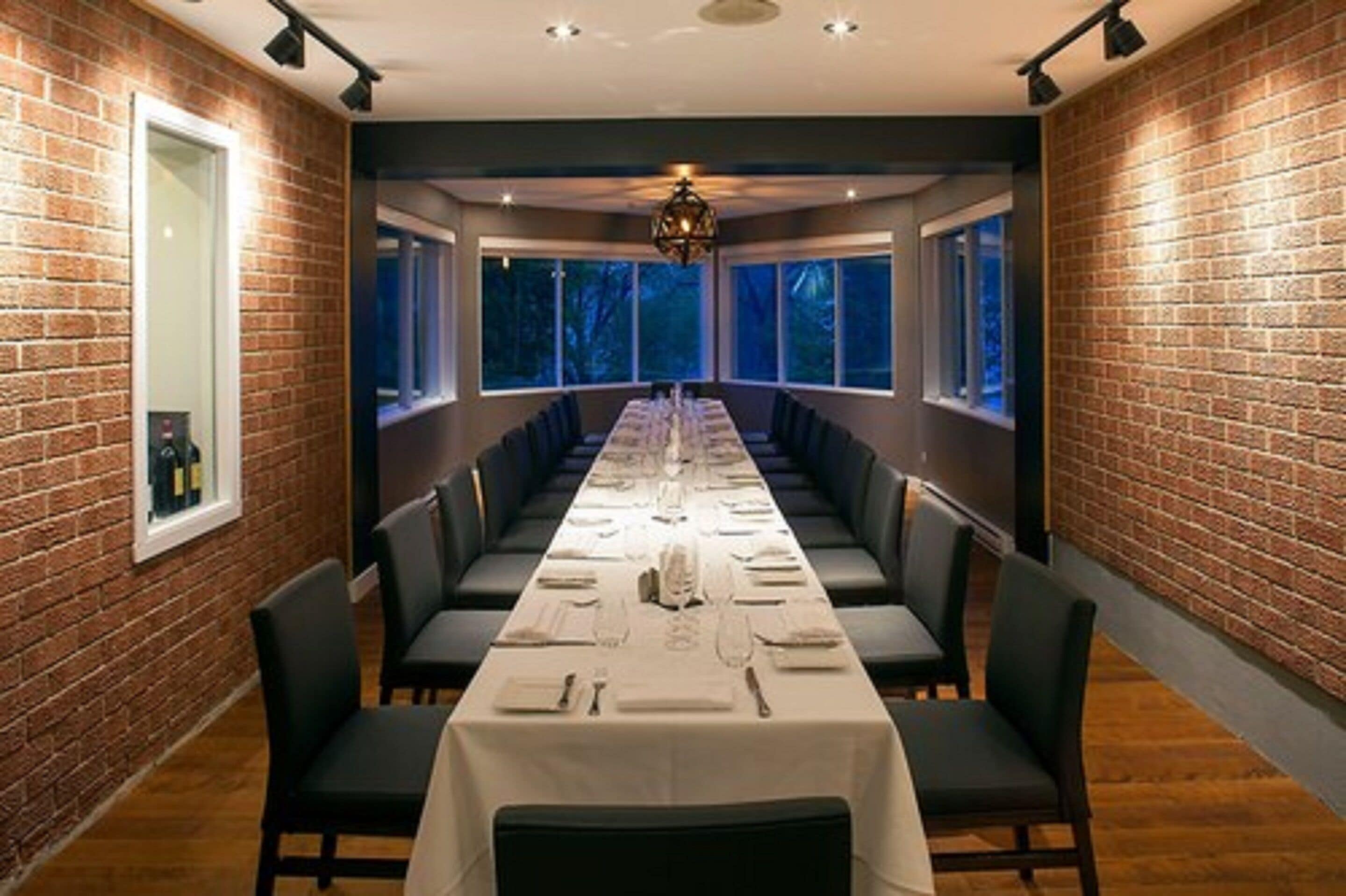 A long dining table set with chairs, large windows, and brick walls.