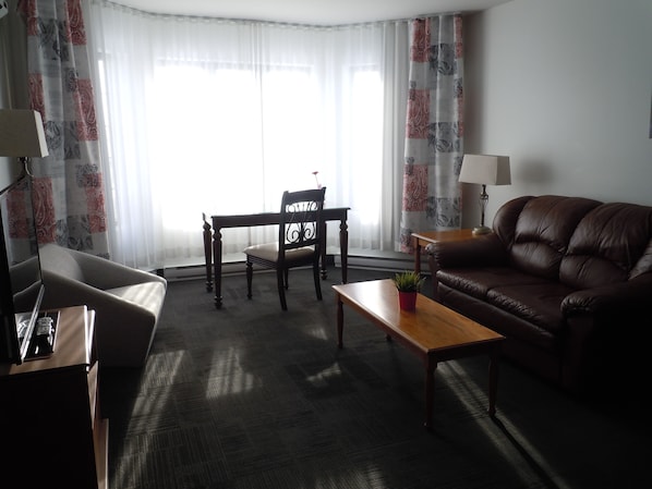 A living room with a brown leather sofa, a wooden coffee table, a wooden chair, and a small table with a lamp.