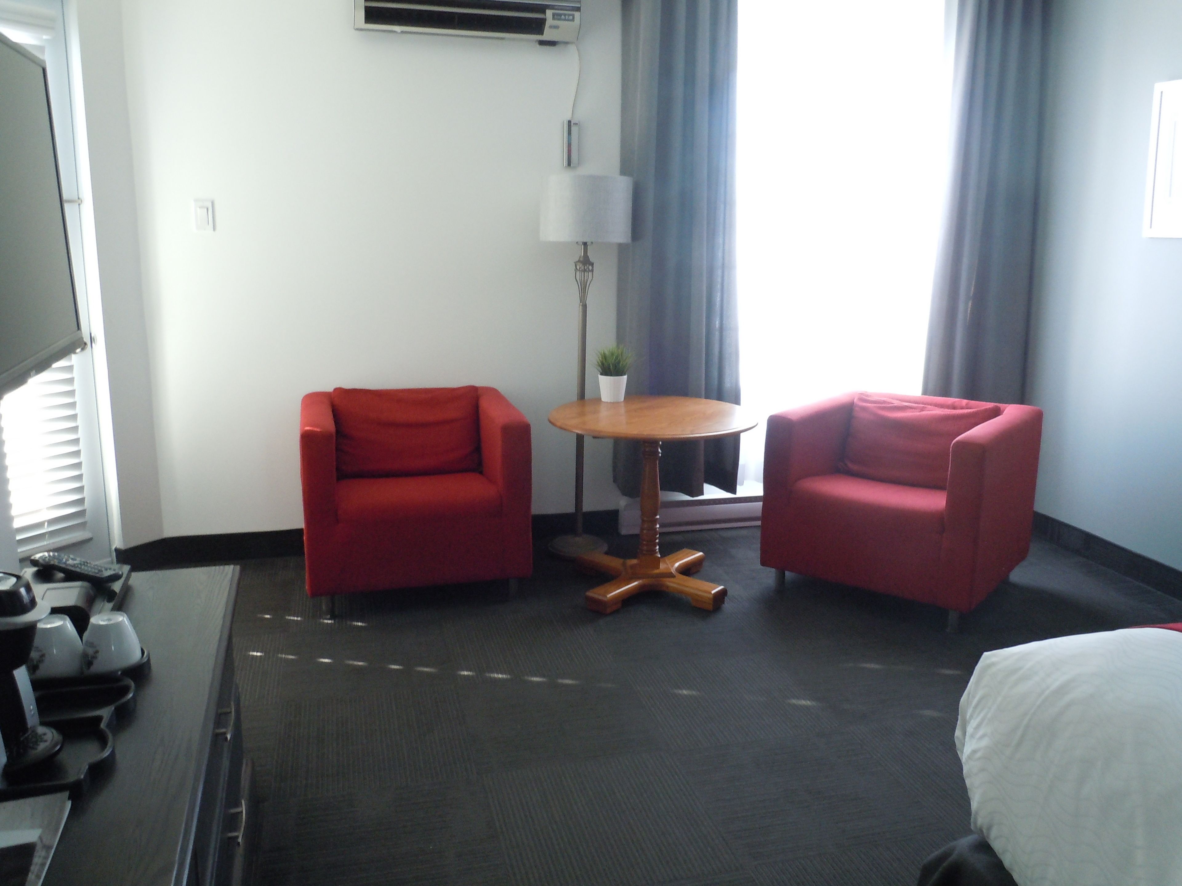 A hotel room with two red armchairs, a small wooden table, a lamp, and a bed.