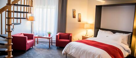 A hotel room with a large bed, a wooden staircase, two red armchairs, a small table with a plant, and a window with curtains.
