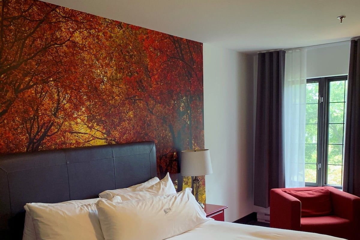 Ein Hotelzimmer mit einem großen Bett, einem roten Sessel, einem Gemälde von Herbstlaub und einem Fenster mit Vorhängen.