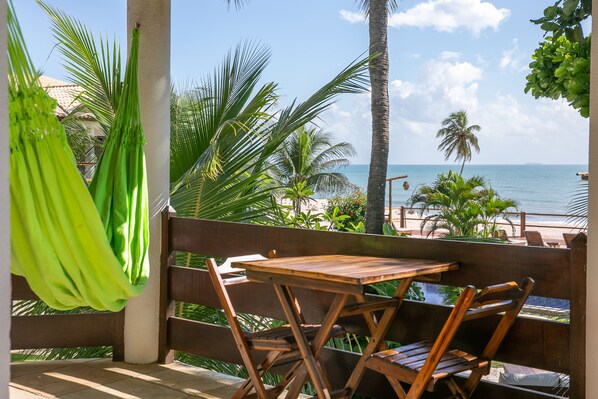 A wooden table and chairs on a balcony overlooking a beach with palm trees and a clear sky.