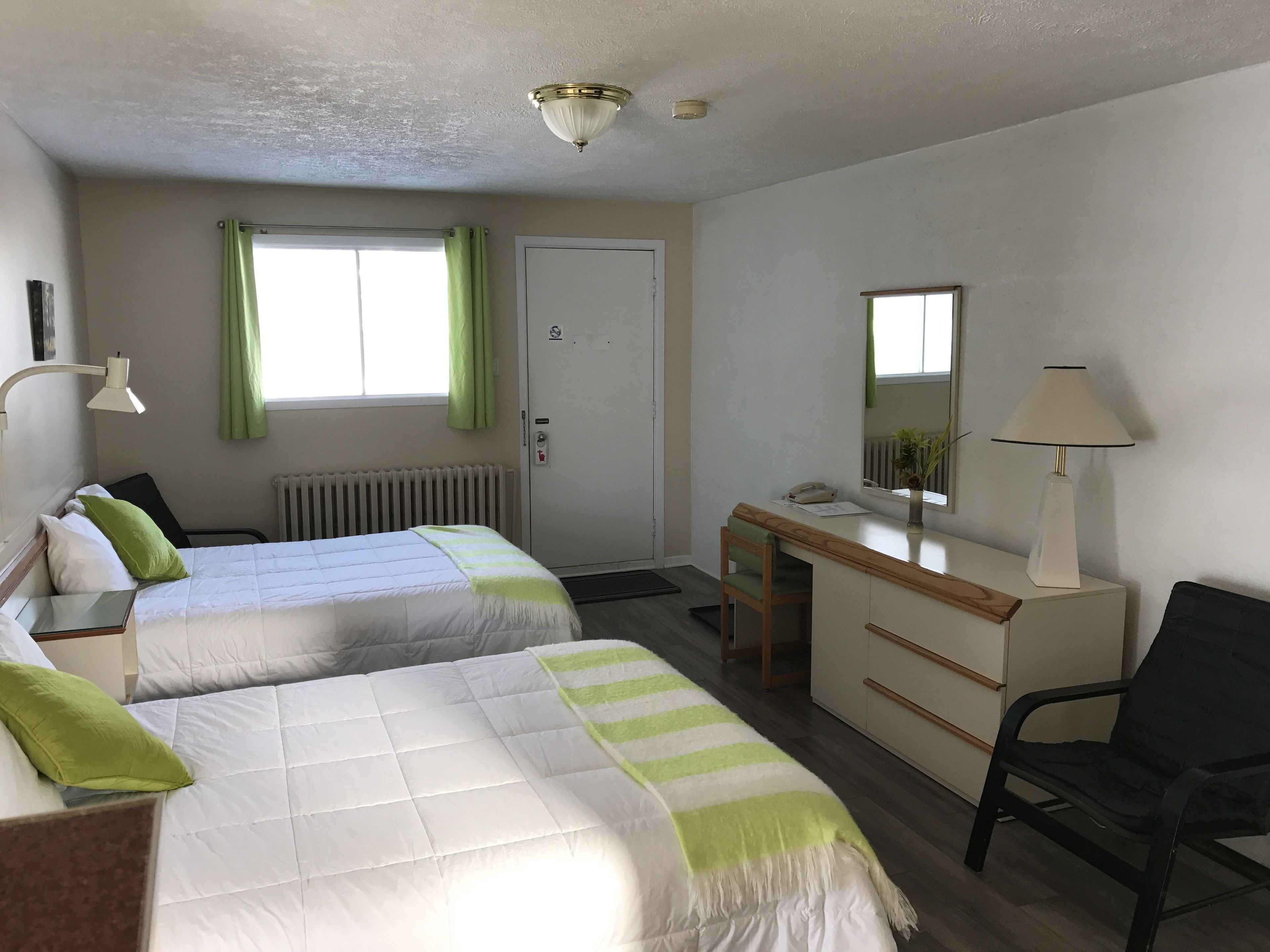 Habitación de hotel con dos camas, un escritorio, una silla, un radiador, una ventana con cortinas e iluminación en el techo.