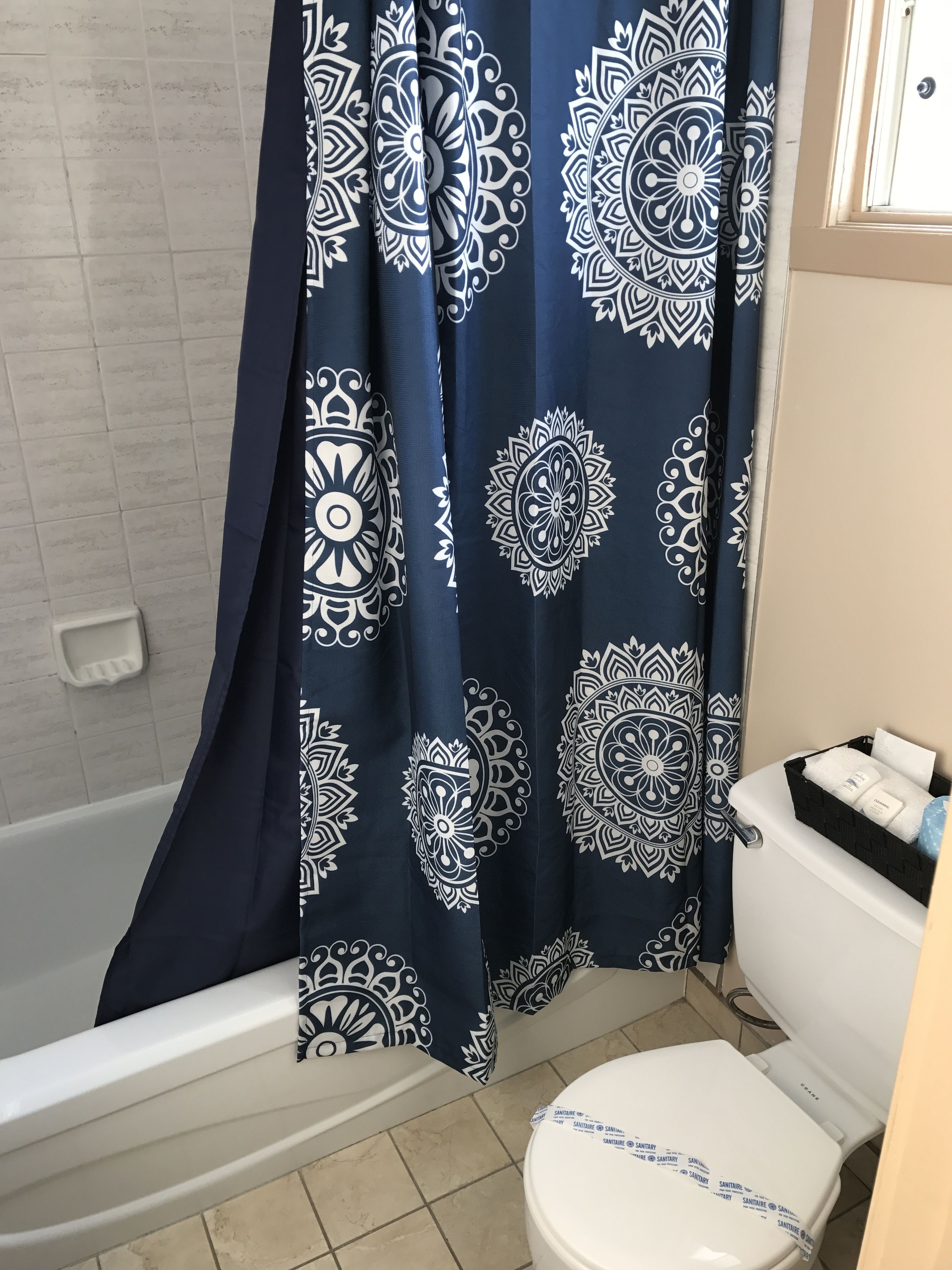 Un baño con inodoro blanco, cortina de ducha blanca y azul estampada y una cesta negra con artículos de aseo.