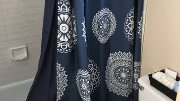 Un baño con inodoro blanco, cortina de ducha blanca y azul estampada y una cesta negra con artículos de aseo.