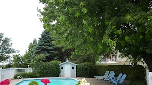 Une piscine entourée d’une clôture blanche, avec un petit abri de jardin et des chaises bleues à l’arrière-plan.