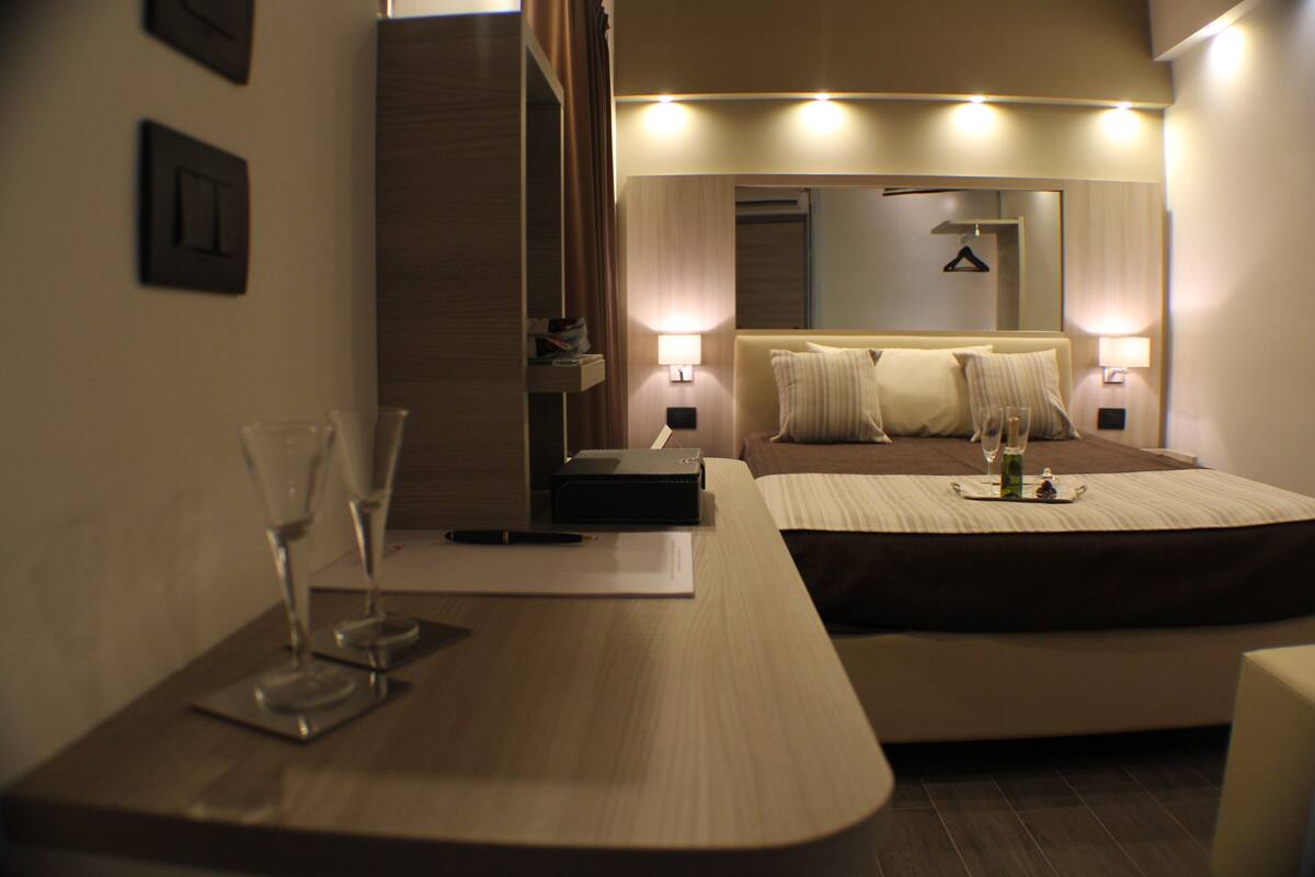 A modern hotel room with a neatly made bed, bedside lamps, a bedside table with a glass of water and a pen, and a mirror above the bed.