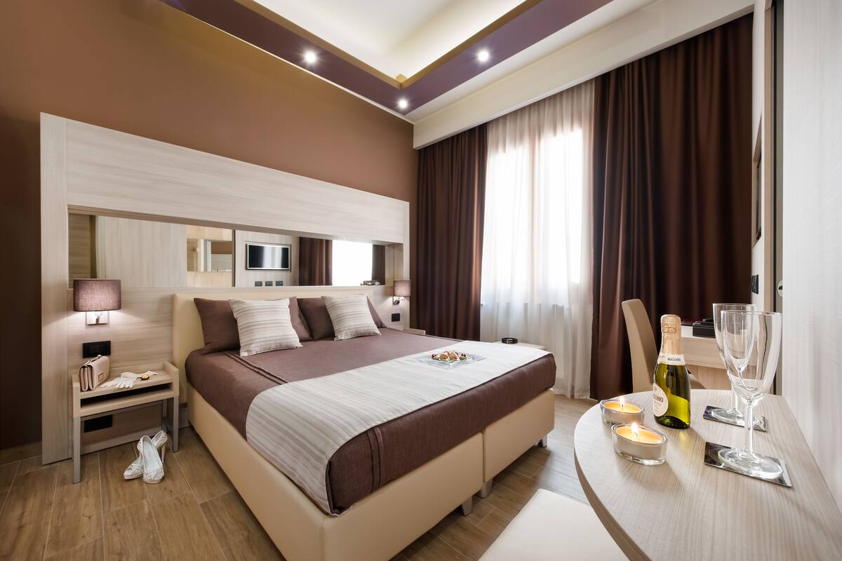 A modern hotel room with a large bed, bedside table, lamp, and a small table with a bottle and glass.