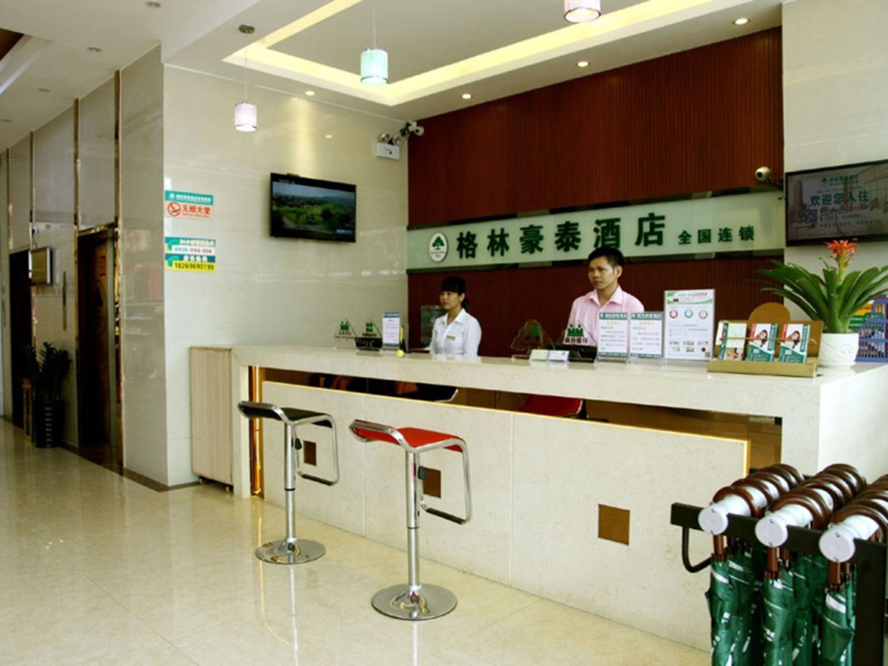 A hotel reception area with a wooden reception desk, two bar stools, a potted plant, and a sign indicating the hotel name.