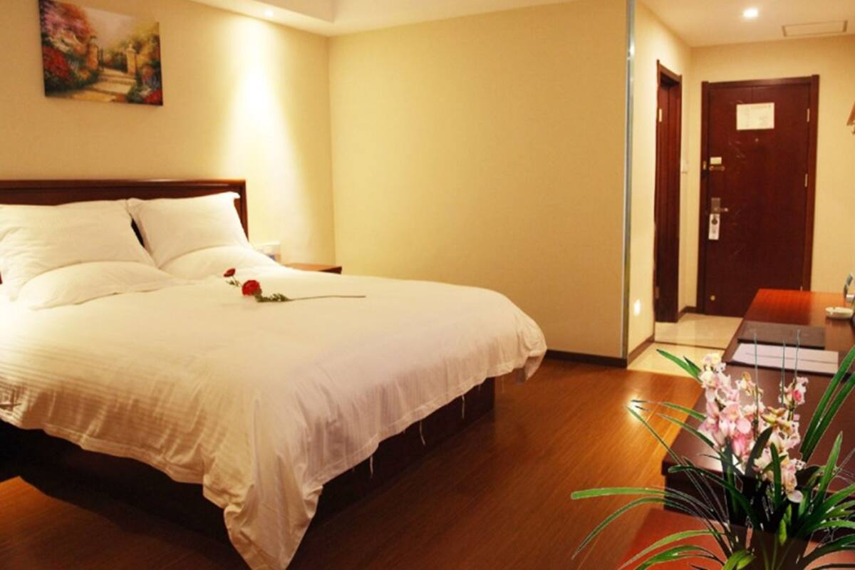 A hotel room with a large bed, a wooden headboard, a painting on the wall, and a desk with a vase of flowers.