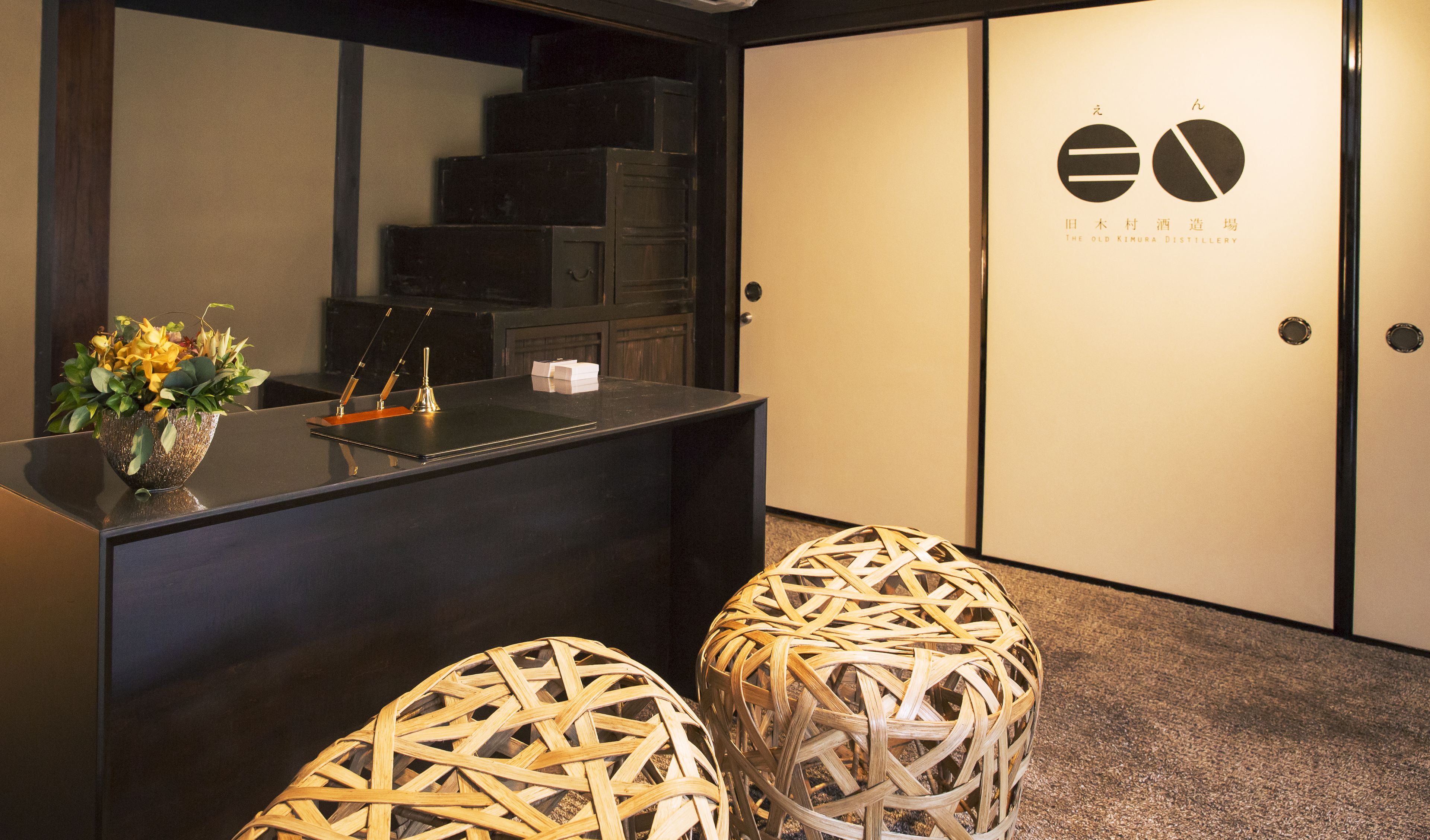 A reception desk with a vase of flowers, a wooden stool, and a sign for The Old Kimura Distillery.