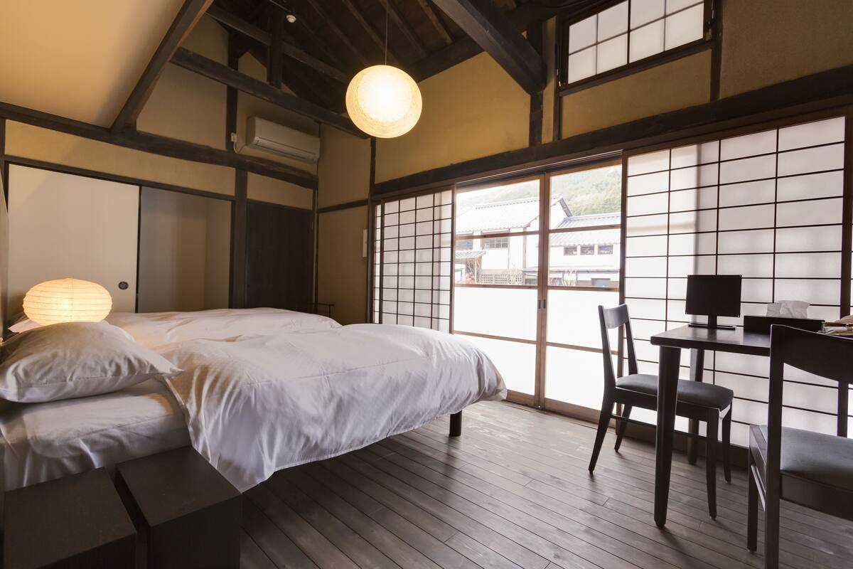 A traditional Japanese-style bedroom with tatami flooring, a bed with white bedding, a desk with a chair, and a large shoji screen door.