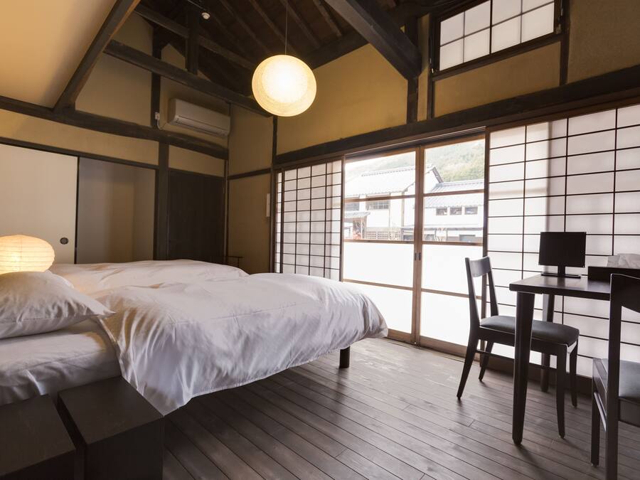 Quarto japonês tradicional com tatames, uma cama com roupa de cama branca, uma mesa com cadeira e um grande portas de papel shoji.