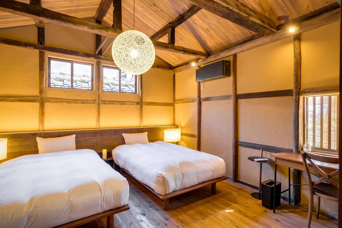 A room with two beds, wooden beams, a hanging light fixture, and a window with blinds.