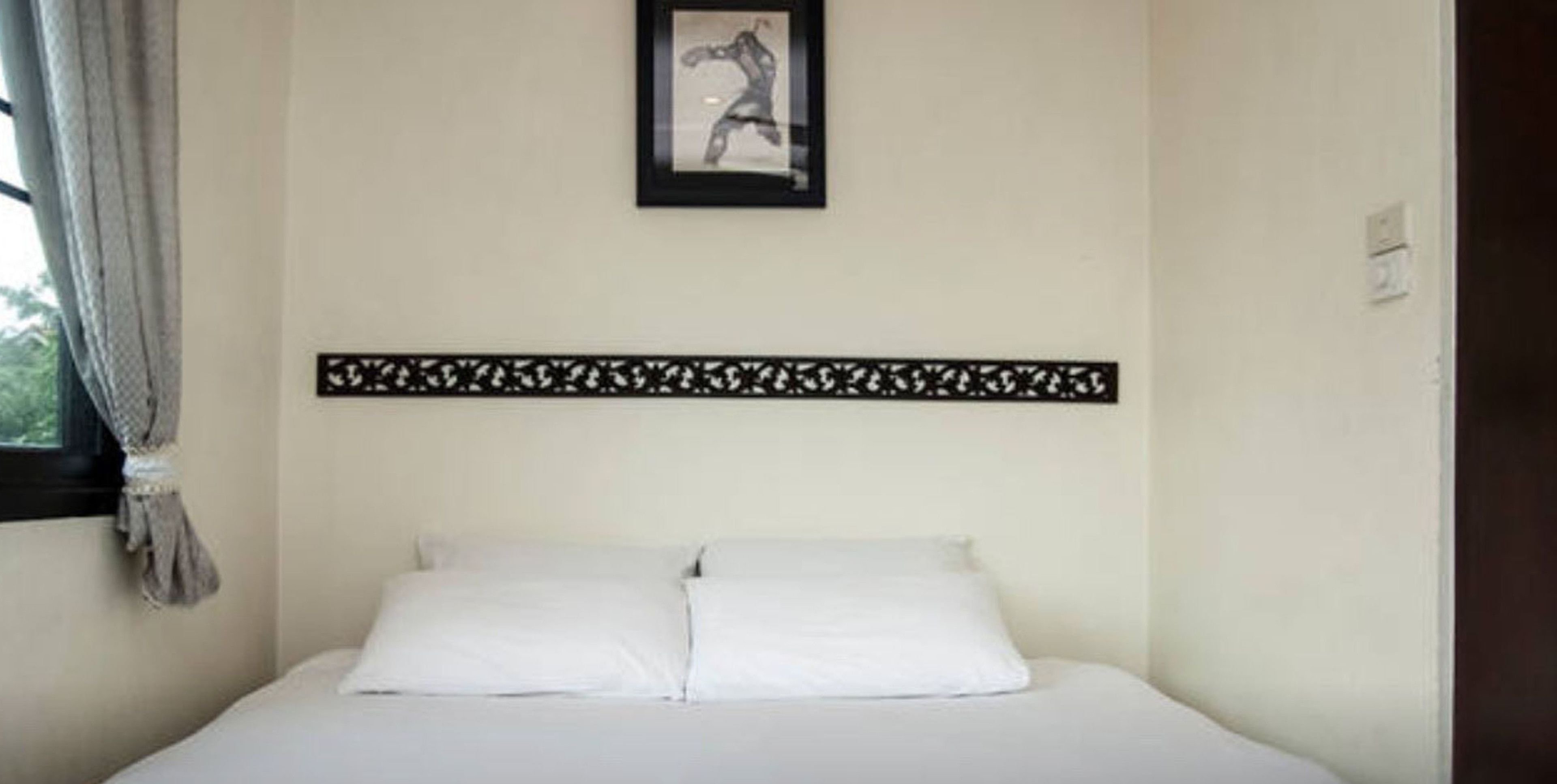 Une chambre d’hôtel avec un lit, une fenêtre avec des rideaux, un tableau encadré au mur et une tête de lit décorative.