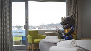 Habitación de hotel con una cama, un sillón verde, una mesa con un peluche y vistas a un paisaje urbano a través de un ventanal grande.