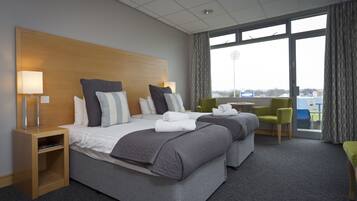 Quarto de hotel com duas camas, uma escrivaninha, uma cadeira e uma janela grande com vista para a cidade.