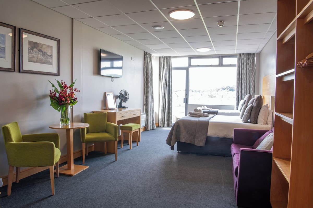 A hotel room with a bed, two chairs, a desk, a TV, and a window with curtains.