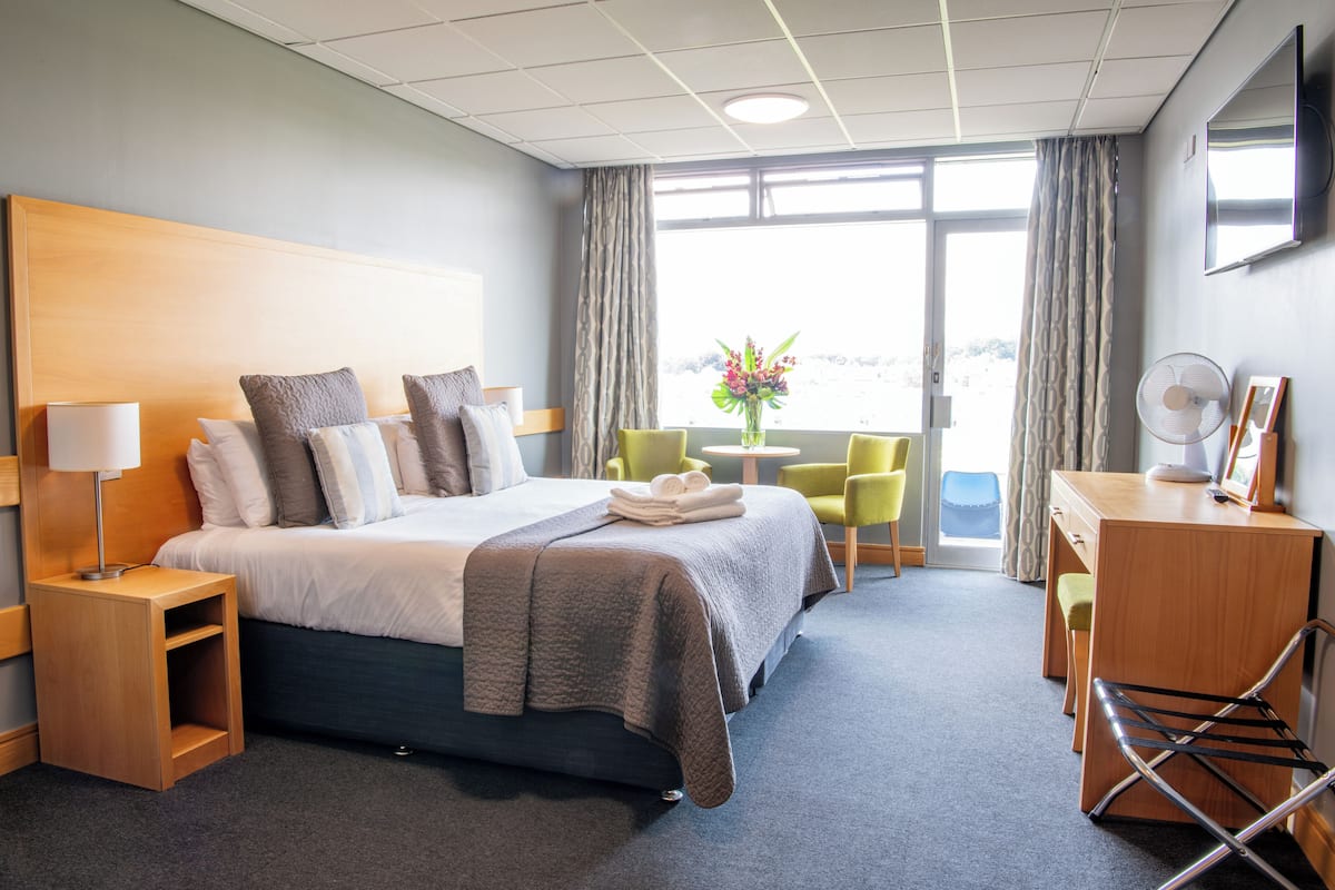 A hotel room with a bed, bedside tables, a desk, a chair, a fan, and a view of the outdoors.