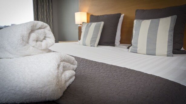 Habitación de hotel con una cama bien hecha, una toalla grande y mullida, y un cabecero con dos almohadas.