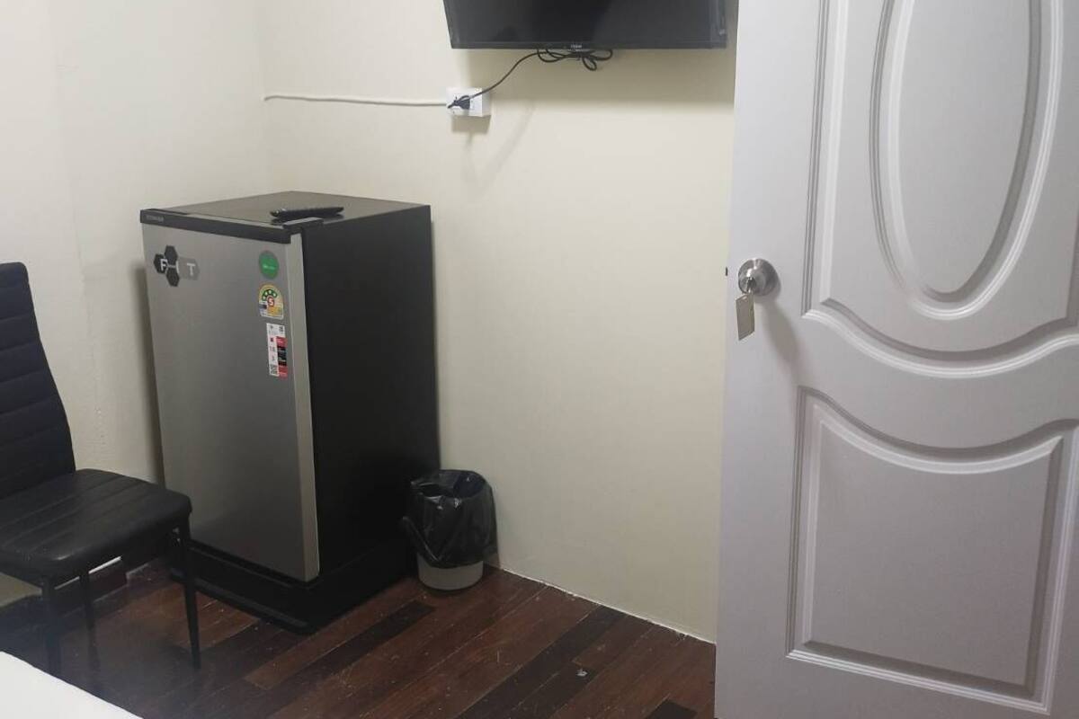 A small room with a bed, a desk, a refrigerator, a trash bin, a television mounted on the wall, and a door with the number 16.