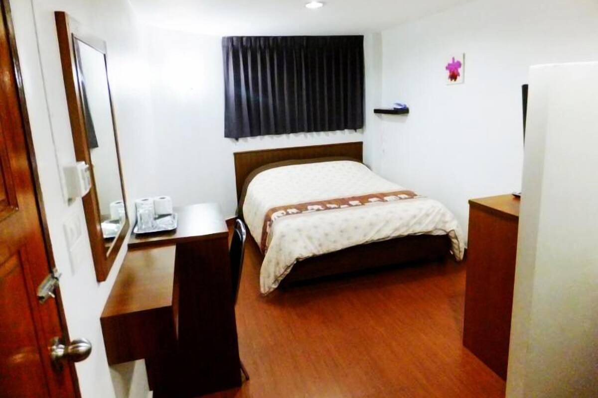 A hotel room with a wooden desk, a bed with a patterned bedspread, a black curtain, and a small decorative item on the wall.