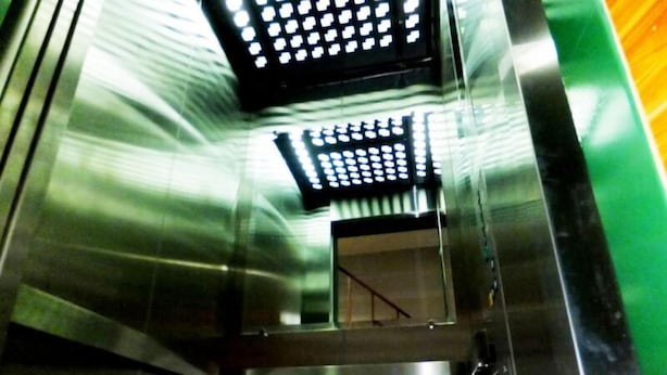 Interior de un ascensor metálico con superficies reflectantes y una pared verde.