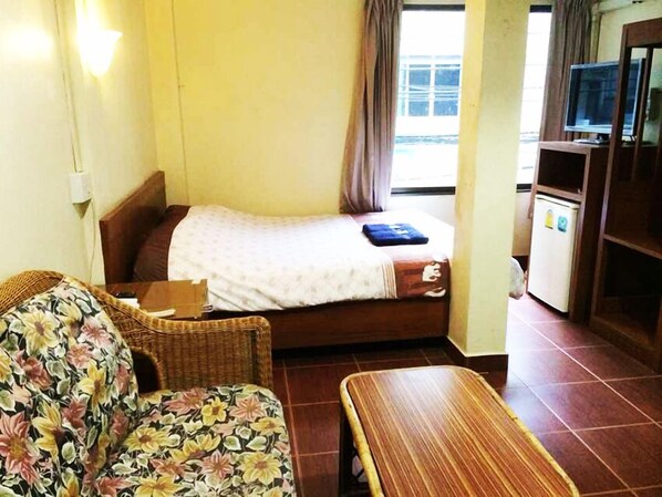 A small, compact room with a single bed, a wooden bench, a wicker chair, and a television on a wooden stand.